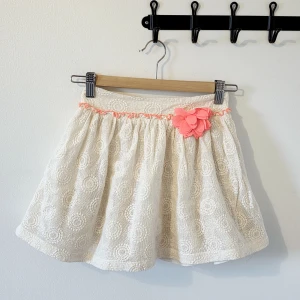 Vit kortkjol med spets och rosa blomma - Supersöt vit kortkjol från Mothercare med broderad spets och cirkelmönster. Kjolen har en ljusrosa dekorationsblomma och matchande rosa kant upptill. Tillverkad i bomull och nylon med polyesterfoder. Perfekt för dig som gillar gulliga detaljer och blommor.