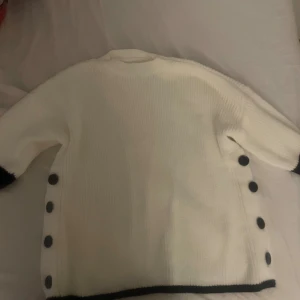 Vit stickad kofta med svarta knappar - Säljer en vit stickad kofta med svarta detaljer och stora svarta knappar längs sidorna. Ärmarna har två svarta ränder och koftan har en ribbad struktur. Perfekt för dig som gillar stilrena och klassiska plagg med en twist.