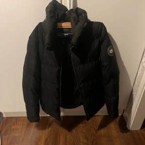 Svart pufferjacka Supply & Demand - Svart pufferjacka från Supply & Demand med huva och dragkedja framtill. Jackan har en patch på ärmen och en rejäl, vadderad känsla som håller dig varm. Perfekt för kalla dagar och har en clean, modern look.