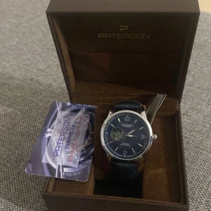 Blå automatisk klocka från Paterson - Snygg blå Paterson automatisk klocka med rund urtavla och silverfärgad boett. Urtavlan har ett mekaniskt fönster och stilrena indexmarkeringar. Armbandet är blått och har en skinnstruktur med synliga sömmar. Levereras i original ask med certifikat.