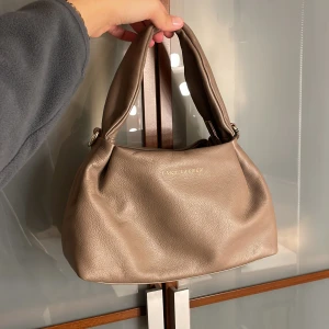 Beige handväska från Lara Lauren - Snygg beige handväska från Lara Lauren i mjukt skinn med stilren, rundad form och breda handtag. Väskan har en enkel design med guldfärgad logga framtill och diskreta metalldetaljer vid handtagen. Perfekt storlek för att få plats med allt du behöver.