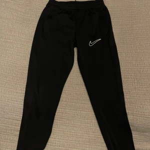 Svarta mjukisbyxor från Nike - Svarta mjukisbyxor från Nike med vit logga på ena benet. Byxorna har elastisk midja och smal passform som sitter snyggt. Perfekta för träning eller chill, tillverkade i mjukt syntetmaterial som känns skönt mot huden.