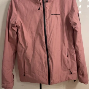 Rosa vindjacka Peak Performance Gore-Tex - Rosa skidjacka från Peak Performance i Gore-Tex-material. Jackan har huva, svarta dragkedjor, två fickor framtill och en ärmficka där du kan lägga liftkortet. Broderad logga på bröstet och ärmen. Perfekt för dagar som är kalla och regniga i skidbacken. Men passar även i pulkabacken och när du åker skridskor.