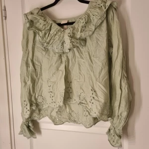 Ljusgrön blus med volang och broderi - Säljer en ljusgrön blus med romantiska volanger och broderade detaljer längs halsringning, ärmslut och nederkant. Blusen har långärmade ärmar med rynk och vacker spetskant. Perfekt för dig som gillar en feminin och bohemisk stil.