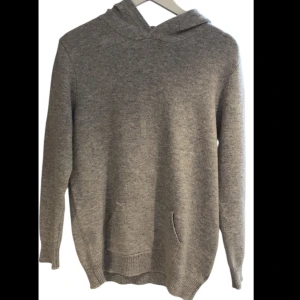 Grå stickad hoodie med huva - Mysig grå stickad hoodie med huva och känguruficka framtill. Tröjan har lång ärm och ribbade muddar vid ärmslut och nederkant. Perfekt för kyliga dagar när du vill vara både bekväm och snygg.