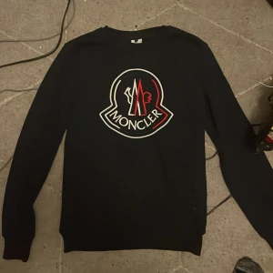 Svart Moncler tröja med stor logga - Svart långärmad tröja från Moncler med stor broderad logga i vitt och rött på bröstet samt en mindre logga på ärmen. Klassisk rund halsringning och ribbade muddar. Perfekt för dig som gillar streetwear och vill sticka ut med en ikonisk design. Storlek L men passar mig som har S
