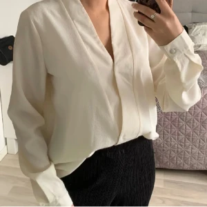 Vit omlottblus från Vila - Stilren vit omlottblus från Vila med v-ringning och långa ärmar. Blusen har en lätt textur i tyget, knappar vid ärmsluten och en avslappnad passform. Perfekt att styla med både jeans och kostymbyxor.