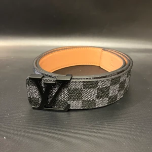 Svart rutig bälte med svart logga - Säljer ett bälte med klassiskt rutigt mönster i svart och grått. Bältet har en bred design med en svart metall logga som spänne. Insidan är i ljusbrunt skinn. Perfekt för dig som vill ha en statement-accessoar.