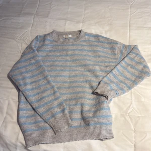 Randig stickad tröja från Gina Tricot - Mysig stickad tröja från Gina Tricot i grått och ljusblått med randigt mönster. Tröjan har rund halsringning, ribbade muddar och är långärmad. Perfekt för lager-på-lager under kyliga dagar.