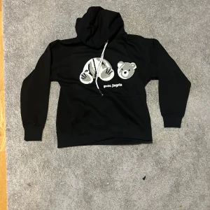 Svart Palm Angels hoodie med nalle - Svart hoodie från Palm Angels med ikoniskt nalletryck på bröstet och vit snörning i huvan. Tröjan har känguruficka och ribbade muddar. Perfekt för dig som gillar streetwear och vill sticka ut med en unik detalj framtill.