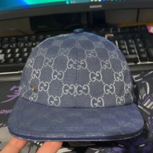 Blå Gucci keps med GG-mönster - Säljer en blå keps från Gucci med klassiskt GG-mönster över hela kepsen. Kepsen har böjd skärm, justerbart spänne bak i blått och är tillverkad i ett slitstarkt tygmaterial. Perfekt för dig som vill sticka ut med en lyxig accessoar.