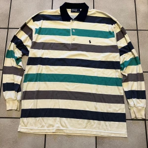 Randig långärmad pikétröja Polo Ralph Lauren - Säljer en klassisk långärmad pikétröja från Polo Ralph Lauren med breda ränder i beige, marinblått, grönt och grått. Tröjan har krage, knäppning framtill och broderad logga på bröstet. Tillverkad i 100% bomull och perfekt för en chill och stilren look.