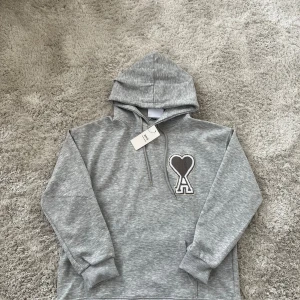 Grå hoodie från Ami Paris  - Säljer en stilren grå hoodie från Ami Paris  med ikoniskt hjärta-A broderi på bröstet. Tröjan har huva med snörning, ribbade muddar och är tillverkad i mjuk bomullsmix. Perfekt för dig som gillar en clean och avslappnad look.
