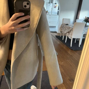 Beige omlottkappa från ONLY - Säljer en beige kappa från ONLY med snygg omlottmodell och bälte i midjan. Jackan har långa ärmar, bred krage och är i ett mjukt material som känns lyxigt. Perfekt för dig som gillar stilrena och enkla plagg.