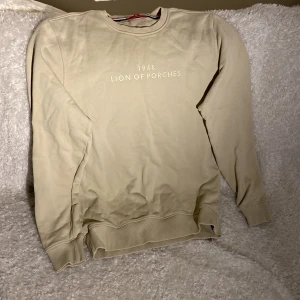 Beige sweatshirt från Lion of Porches - Snygg beige sweatshirt från Lion of Porches med tryck '1948 LION OF PORCHES' på bröstet. Tröjan har rund hals, ribbade muddar och är långärmad. Perfekt för dig som gillar stilrena och enkla plagg med diskret branding. Medium 