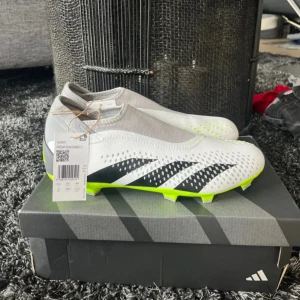 Adidas Predator Accuracy 3 fotbollsskor - Säljer ett par Adidas Predator Accuracy 3 fotbollsskor i vitt med svarta detaljer och neongul sula. Skorna har en strumpliknande passform utan snörning och är tillverkade i syntetmaterial. Perfekta för dig som vill sticka ut på planen.