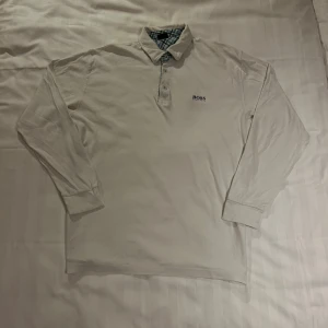 Vit långärmad Piké från Hugo Boss - Vit långärmad Piké från Hugo Boss med klassisk krage och knappar framtill. Insidan av kragen har ett rutigt mönster i grönt, blått och vitt. Skjortan är i mjuk bomull och har diskret Boss-logga på bröstet.