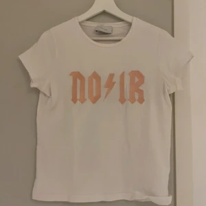 Vit t-shirt från Neo Noir - Vit t-shirt från Neo Noir med korta ärmar och rund halsringning. Framsidan har ett coolt persikofärgat NOIR-tryck med blixtdetalj i mitten. 