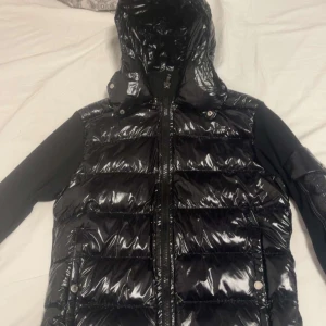 Svart Moncler cardigan - Svart pufferjacka från Moncler med glansig finish och huva. Jackan har Moncler-logga på ärmen, dragkedja framtill och tryckknappar på fickorna. Materialet är syntet och polyester, perfekt för kalla dagar. Snygg och modern look med quiltade paneler och långärmade svarta ärmar.