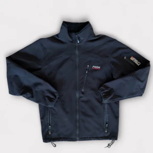 Svart softshell Peak Performance - Svart vindjacka från Peak Performance med broderad logga på bröstet och ärmen. Jackan har hög krage, tre dragkedjefickor och grått fleecefoder på insidan. Perfekt för dig som gillar sportig stil och vill ha något funktionellt och snyggt.