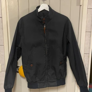 Svart vindjacka från Fred Perry S - Stilren svart vindjacka från Fred Perry med klassisk krage, dragkedja framtill och snygga knappar vid halsen. Jackan har två fickor med knappstängning och diskret broderad logga på bröstet. Perfekt för dig som gillar enkel och tidlös design.