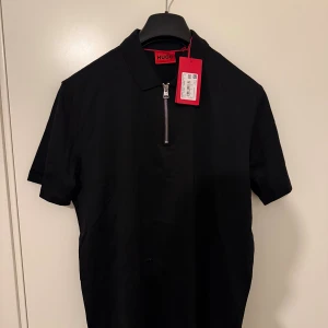Svart t-shirt med dragkedja från Hugo - Svart t-shirt från Hugo med krage och snygg half zip-dragkedja framtill. Klassisk passform och korta ärmar, perfekt för en clean och stilren look. Tillverkad i mjuk bomull för maximal komfort. Helt ny med tagg 
