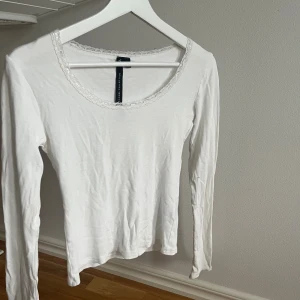 Vit långärmad topp med spetskant - Säljer en vit långärmad topp. Toppen har en rundad halsringning med fin spetskant som ger en söt touch. Materialet känns mjukt och lätt, perfekt för dig som gillar stilrena och enkla plagg. Passar dig som vill ha en basic topp med lite extra detalj.