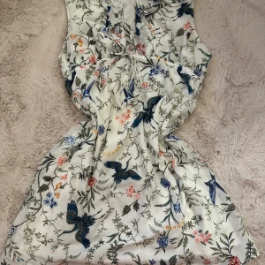 Vit blommig kortklänning med fågelmotiv - Superfin vit kortklänning med blommigt och fågelmönster i blått, grönt och rosa. Klänningen har omlottvolanger vid halsen och ett knytband i midjan för extra snygg passform. Perfekt för dig som gillar romantiska och detaljerade prints.