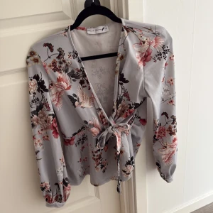 Blommig omlottblus från Black Story -  Ser jättestor ut! Är den inte! Vanlig i storlek Supersnygg blommig omlottblus från Black Story i ljusgrått med rosa och beige blommor samt fågelmotiv. Blusen har långa ärmar med lätt puff och knyt framtill för en extra fin siluett. Perfekt för dig som gillar romantiska detaljer och v-ringad stil.