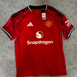 Manchester United Cunha 10 Adidas tröja - Röd Manchester United matchtröja från Adidas med vita detaljer, svart-vita muddar och klubbmärke på bröstet. Tryck med Snapdragon och nummer 10 Cunha på ryggen. Tillverkad i lätt och ventilerande material, perfekt för fotboll eller supporterliv.