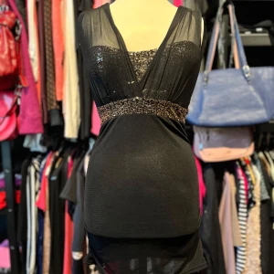 Svart glittrig kortklänning/topp - Svart kortklänning/topp med glittriga paljetter över bysten och midjan. Klänningen har ett snyggt omlottliv med transparent mesh upptill och en figurnära passform. Axelbandslös design framtill och v-ringad rygg ger en cool och festlig look. Några paljetter är borta framtill (se bild 3), men inget som syns när mesh tyget är uppe.