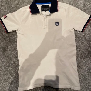 Vit Sail Racing pikétröja USA 77 - Vit pikétröja från Sail Racing med mörkblå krage och röda detaljer. Broderad USA 77-text på ryggen och amerikansk flagga på ärmen. Klassisk passform och korta ärmar, tillverkad i bomull. Perfekt för dig som gillar sportig och stilren look.