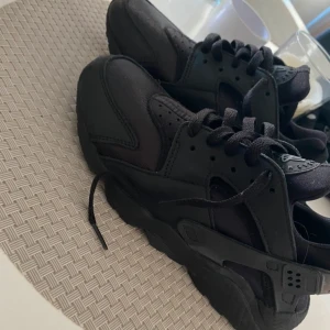 Svarta Nike Huarache sneakers - Säljer ett par helsvarta Nike Huarache sneakers med klassisk chunky sula och snygga detaljer i både textil och syntet. Skorna har snörning och rund tå, samt den ikoniska Huarache-loggan på plösen och bakre hälremmen.  Dem är använda 2 gånger så i ett bra skick! 