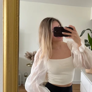 Vit croppad blus med puffärmar - Säljer en vit croppad blus med transparenta puffärmar i organza och fyrkantig urringning. Toppen har en slim passform och är riktigt snygg till både jeans och kjol. Ärmarna är långa och har volangdetaljer vid mudden för extra statement.