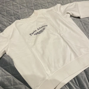 Vit sweatshirt Paris Atelier H&M XS - Vit sweatshirt från H&M i storlek XS med broderad text 'Paris Atelier' och adress på bröstet. Tröjan har rund hals, ribbade muddar och lång ärm. Mjuk insida och klassisk passform, perfekt för chill dagar. Ganska mycket ludd längst armar.