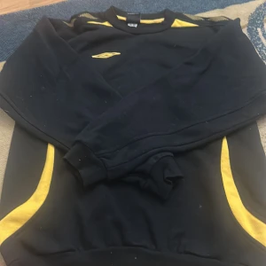 Svart och gul Umbro sweatshirt - Cool svart sweatshirt från Umbro med gula detaljer på axlar och sidor samt broderad logga på bröstet. Tröjan har rund halsringning och långa ärmar, perfekt för dig som gillar sportig stil.