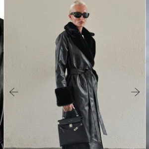 Svart lång skinnkappa med fuskpäls - Lång svart kappa i skinnimitation från NA-KD med bred krage och muddar i mjuk fuskpäls. Kappan har bälte i midjan, knappar framtill och är även vadderad