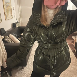 Grön jacka med bälte - Säljer en mörkgrön, glansig pufferjacka med luva och bälte i midjan som man kan välja att ha eller inte. Jackan har en cool unik grön skiftande färg 