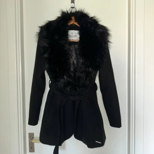 Svart kappa med fuskpäls från Chiara Forthi - Stilren svart kappa från Chiara Forthi Milano med bred krage i fuskpäls och knytskärp i midjan. Jackan har lång ärm och två fickor framtill. Perfekt för dig som vill ha en elegant look med extra edge.