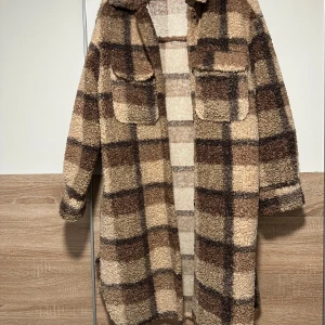 Rutig teddyjacka i bruna toner - Mysig teddyjacka med rutigt mönster i beige, brunt och mörkbrunt. Jackan har två stora bröstfickor med lock, lång ärm och en oversized passform. Perfekt för kalla dagar när du vill ha både stil och komfort. Har även slits på sidorna, endast använd ett fåtal gånger.