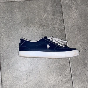 Mörkblå sneakers från Polo Ralph Lauren - Snygga mörkblå sneakers från Polo Ralph Lauren med vit logga på sidan och vit sula. Skorna har rund tå, snörning med vita skosnören och är tillverkade i syntetmaterial med textilfoder. Perfekt för dig som gillar klassisk och sportig stil. Skorna är lite slitna men funkar helt utmärkt. Skriv om du undrar något😃