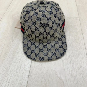 Gucci baseball keps Grå/marinblå - Snygg keps från Gucci i grå med mörkblått GG-logomönster över hela kepsen. Klassisk form med böjd skärm och justerbar rem baktill. Materialet är bomull och polyester, med insida i marinblått tyg. Röd och blå rand på sidan ger extra lyxig känsla.