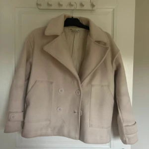 Beige skepparkavaj från Designbysu - Stilren beige skepparkavaj från Designbysi med dubbelknäppning, bred krage och stora fickor framtill. Jackan har en rak, boxig passform och är tillverkad i ett mjukt ullmaterial. Perfekt för dig som vill ha en klassisk look med modern twist.