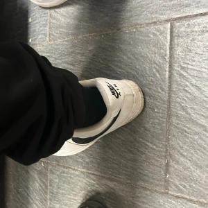 Nike Air Force 1 vita sneakers - Säljer ett par klassiska Nike Air Force 1 sneakers i vitt skinn med svarta swoosh-detaljer. Skorna har rund tå, platt sula och snörning framtill. Perfekt för dig som gillar streetstyle och vill ha en tidlös sneaker med snygg kontrast mellan vitt och svart.