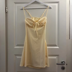 Ljusgul kort klänning med öppen rygg - Supersöt ljusgul kort klänning med smala axelband och söt knytning framtill. Klänningen har en snygg öppen rygg och lätt utsvängd kjol. Aldrig använd!