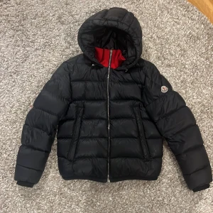 Svart Moncler pufferjacka med huva - Svart pufferjacka från Moncler med huva och röd insida vid halsen. Jackan har quiltad design, dragkedja framtill och två sidofickor. Klassisk Moncler-logga på ärmen. Perfekt för kalla dagar och riktigt snygg streetstil.