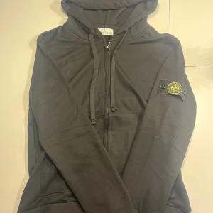 Svart hoodie från Stone Island M - Svart hoodie från Stone Island med klassisk patch på vänster ärm. Tröjan har huva med snörning, dragkedja framtill och ribbade muddar. Materialet är mjuk bomull som känns skön mot huden. Perfekt för dig som gillar streetwear och vill ha en clean look. Hör av dig vid minsta fundering!