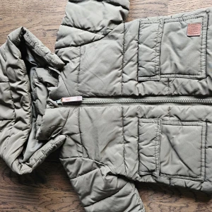 Olivgrön jackan stl 74 - Olivgrön, vadderad jacka med huva från Marie Serneholt. Jackan har två fickor framtill och muddar i ärmslut. Muddarna är lite noppriga och det finns en diffus liten fläck på jackan.