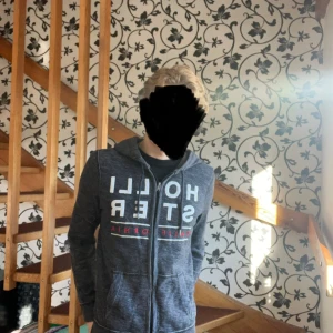 Mörkgrå Hollister hoodie med dragkedja - Snygg mörkgrå hoodie från Hollister med stor vit logotyp och röd text på framsidan. Kom gärna med rimliga bud! Modellen är 181cm/70kg