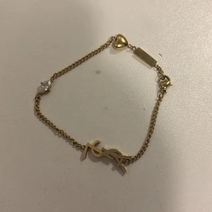 Guldigt armband med hjärtdetalj - Snyggt armband i guldton  med en liten hjärtdetalj och en glittrande sten. Kedjan är tunn och elegant, perfekt för att ge din outfit en lyxig touch. Lås med karbinhake för enkel användning. Inte äkta